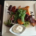 Grillspieß mit Hühnchen, Schweinefleisch und Würstchen auf Kartoffelspalten, buntem Gemüse und Salat, dazu Aioli